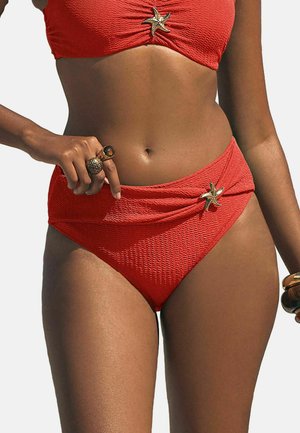 Femeie purtând un costum de baie texturat roșu, decorat cu stele de mare pe top și chiloți cu talie înaltă, arătând mijlocul și mâna cu inele.