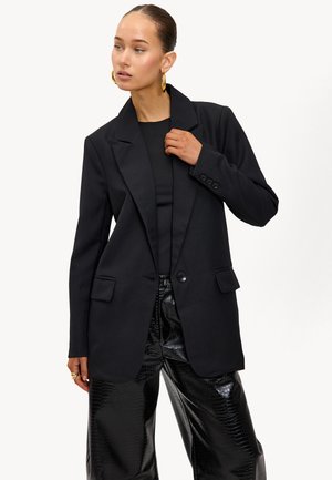 Blazer noir ajusté avec revers crantés, fermeture à un bouton et poches devant, associé à un pantalon noir vernissé avec motif crocodile.