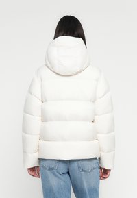 Chaqueta de plumas blanca con capucha, con un diseño acolchado y mangas anchas. La tela parece suave y ligeramente brillante.