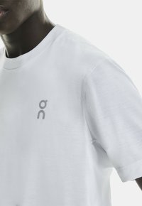 T-shirt en coton blanc avec un col arrondi, des manches courtes et un petit logo gris sur la poitrine. Texture lisse, coupe décontractée.
