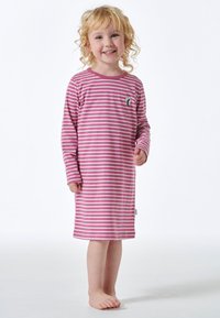 Uśmiechnięte małe dziecko z kręconymi blond włosami, ubrany w różowo-biały paskowany sukienkę z długim rękawem, stoi boso na białym tle.