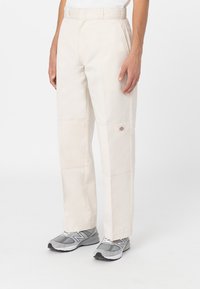 Pantalones de algodón beige con corte recto, con detalles de costura visibles y un pequeño logo en la parte inferior de la pierna; combinados con zapatillas deportivas grises.