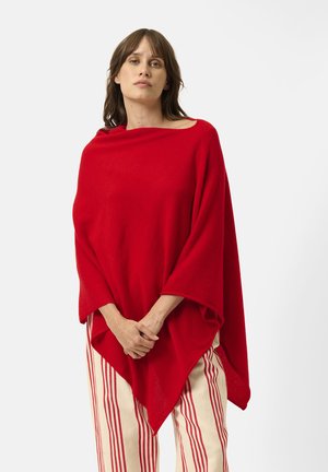 Roter Poncho mit weiten Ärmeln, aus weichem Stoff gefertigt. Mit einem Bootsausschnitt und einem drapierten Design, kombiniert mit gestreiften Creme- und roten Hosen.