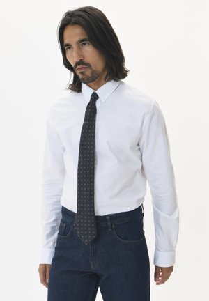 Camisa blanca de botones con mangas largas, combinada con unos pantalones de mezclilla oscuros. La corbata delgada tiene un patrón sutil en colores oscuros.