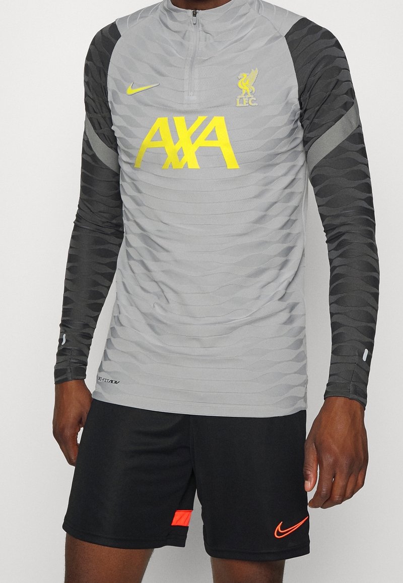 Haut de sport gris à manches longues avec corps texturé, logo jaune et manches noires à motifs, associé à un short noir avec un accent orange.