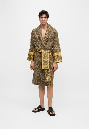 Uomo che indossa un accappatoio marrone con motivo del logo Versace, rifiniture dorate ornate su maniche e cintura, e sandali neri, in piedi su uno sfondo bianco semplice.