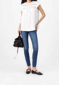 Blusa bianca con pizzo orizzontale decorativo, jeans skinny blu e ballerine nere con tagli circolari, mentre tiene una borsa a sacco nera con motivo floreale.