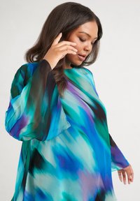 Flowy blouse in sheer fabric featuring a blue-green and purple abstract pattern. Long bell sleeves, round neckline, and relaxed fit.