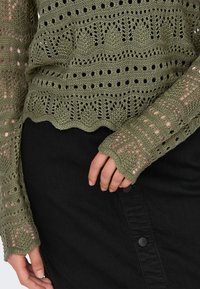 Oliwkowy, dzianinowy sweter z wyrafinowanymi wzorami ażurowymi i falbankowym dołem; zestawiony z czarną spódnicą dżinsową z detalami w postaci guzików.