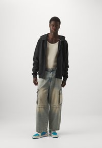 Sort zip-up hoodie, creme tank top, affarvede brede cargo jeans med sidelommer og turkise sneakers med lilla accenter.