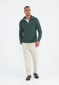 Zelený pletený svetr s límcem na čtvrtinový zip, s ribbed texturou, nošený přes bílou košili, doplněný béžovými kalhotami a hnědými botami.