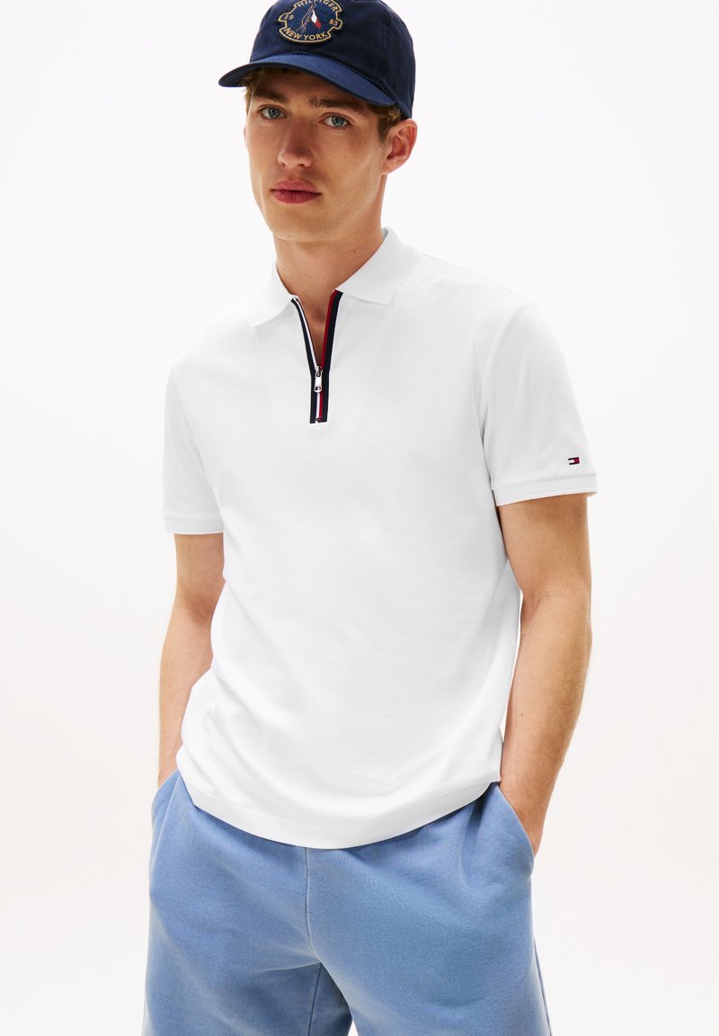 Jonge man in een witte polo met ritssluiting, lichtblauwe shorts en een marineblauwe pet met geborduurd logo, met de handen in de zakken, neutrale achtergrond.