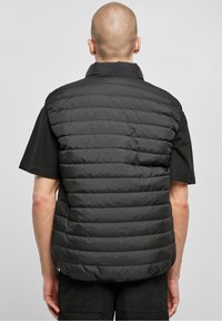 Černá péřová vesta s horizontálním prošíváním, vysokým límcem a uvolněným střihem, vrstvená přes černé tričko s krátkým rukávem.