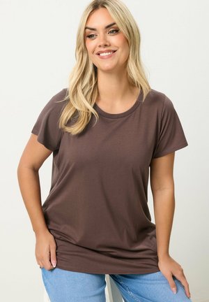 Donna bionda che sorride, indossa una maglietta marrone a maniche corte e jeans azzurri, in piedi con le mani appoggiate sui fianchi.