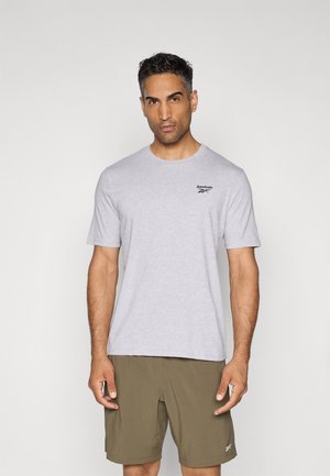 T-shirt en coton gris à manches courtes et col rond, avec un logo Reebok noir sur la poitrine gauche. Porté avec un short vert.