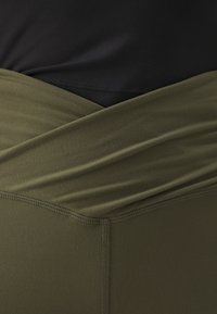 Μαύρο τοπ με σταυρωτό ελαστικό λαιμόκομμα σε ελιά, φτιαγμένο από μαλακό, ελαστικό ύφασμα. Ορατές ραφές κατά μήκος των άκρων, εφαρμοστή σχεδίαση.