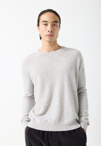 Noor mees tumedate juustega, mis on tagumikku seotud, kannab helehallist kampsunit ja tumedaid pükse, seisab käed taskutes valge tausta ees.