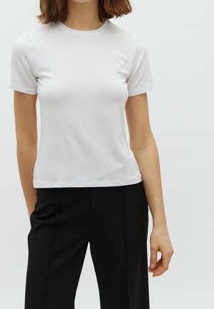 Mujer con camiseta blanca lisa de manga corta y pantalones negros, de pie frente a un fondo blanco, recortada a la altura del cuello.