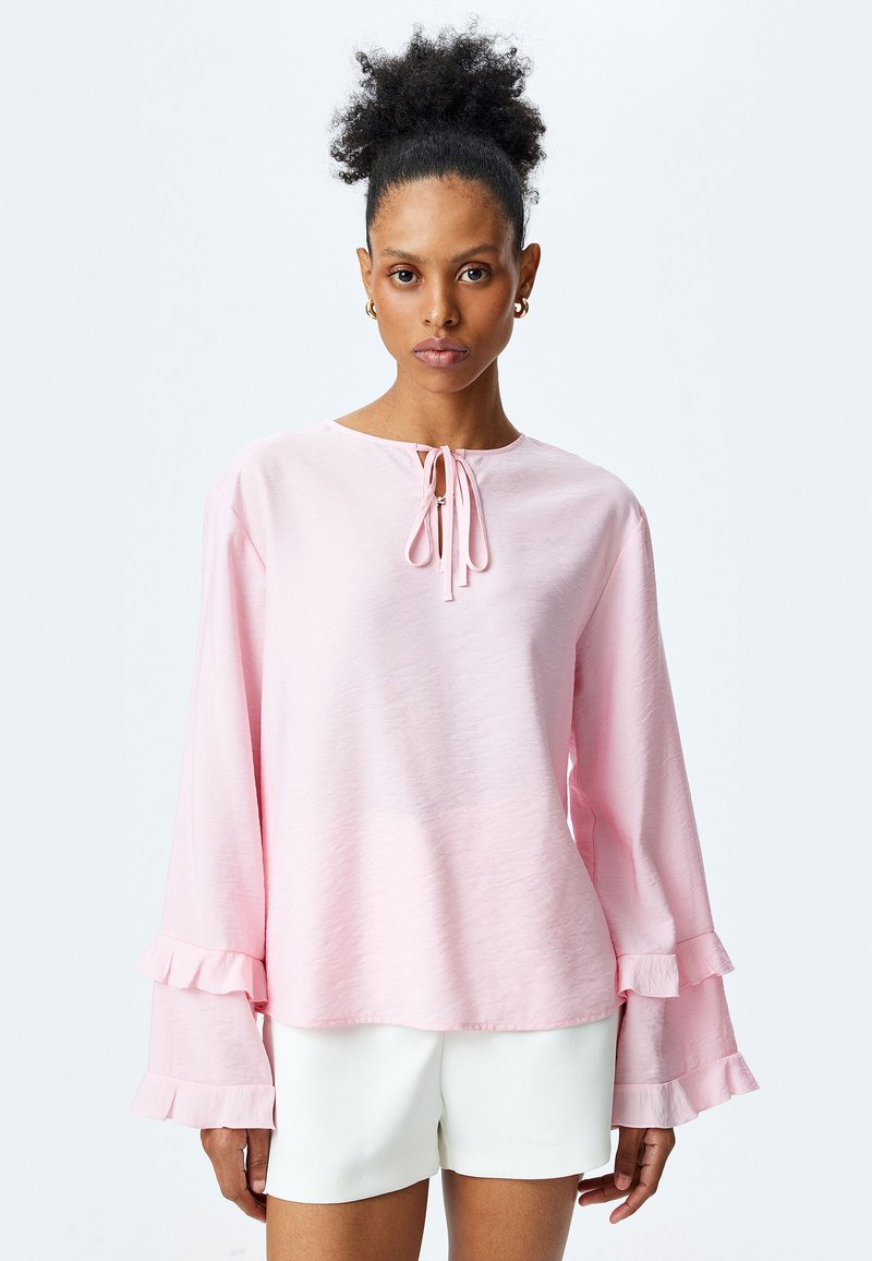 Blusa a maniche lunghe rosa chiaro con scollo a cravatta e polsini arricciati, realizzata in tessuto morbido. Presenta una vestibilità rilassata e una consistenza liscia.