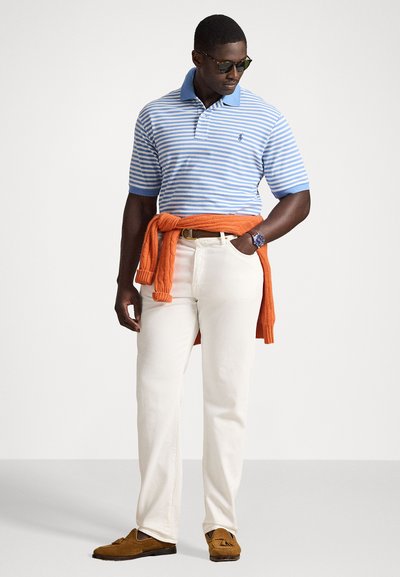 Camisa polo riscada em azul e branco com colarinho azul, calças brancas e mocassins de camurça castanhos; suéter laranja atado à cintura.