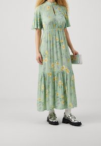 Grünes Blumenkleid mit kurzen, ausgestellten Ärmeln, Schlüsselloch-Ausschnitt und gestuftem Saum. Kombiniert mit schwarzen Plateauschuhen und einer metallischen Clutch.