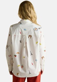 Woman with long blonde hair wearing a white shirt featuring colorful Paris-themed illustrations, standing against a plain background.