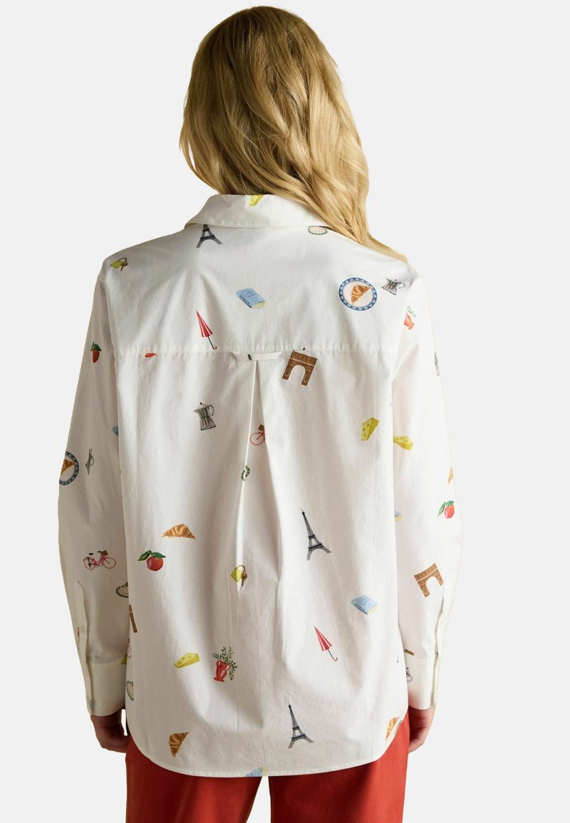 Woman with long blonde hair wearing a white shirt featuring colorful Paris-themed illustrations, standing against a plain background.