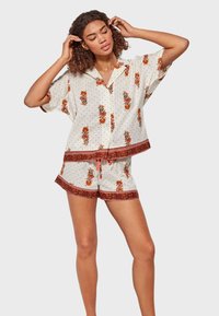 Conjunto de pijama corto impreso en color crema claro, con patrones florales y lunares, y borde rojo en los pantalones cortos y la parte superior. Diseño de manga corta.