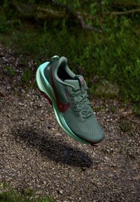 Scarpa da corsa Nike verde e turchese con logo rosso che galleggia sopra un sentiero di ghiaia circondato dall'erba.