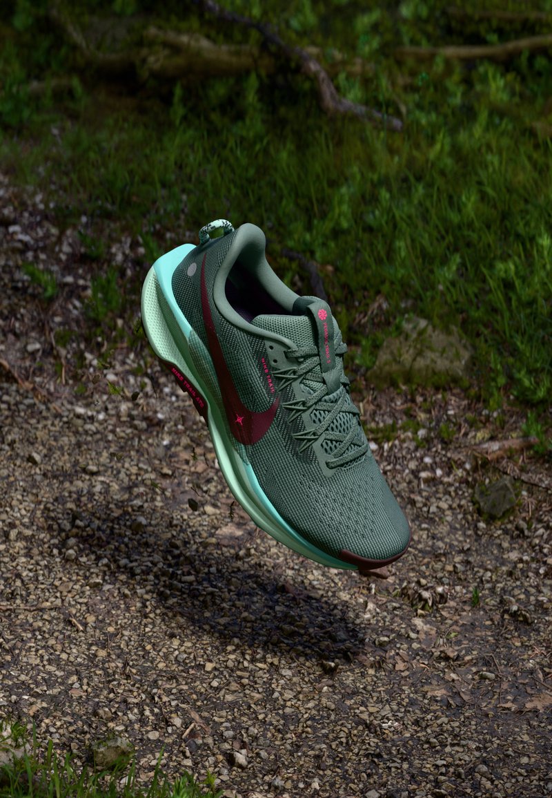 Scarpa da corsa Nike verde e turchese con logo rosso che galleggia sopra un sentiero di ghiaia circondato dall'erba.