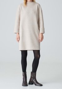 Robe-pull oversize beige clair à manches longues et col rond, associée à des collants noirs et des bottines marron.