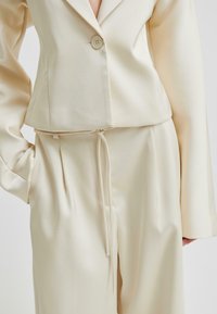 Kvinna klädd i en skräddarsydd kostym i krämfärg med enknappsjacka och byxor med dragsko, med handen i fickan, stående mot en enfärgad bakgrund.