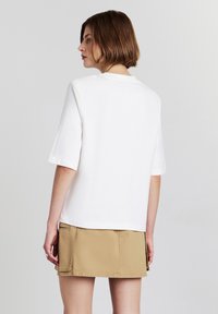 T-shirt blanc à manches courtes avec une coupe décontractée. Le modèle porte une jupe beige, de dos, mettant en valeur le design au dos du t-shirt.