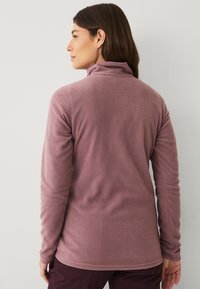 Fleece-trøje i lys mauve med høj krave, lange ærmer og en blød overflade, der har diskrete sydetaljer ved kanten og manchetterne.