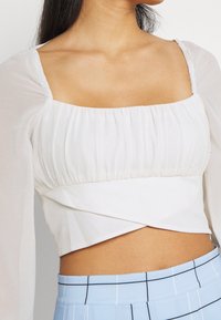 Haut court blanc avec manches bouffantes et encolure carrée ; tissu plissé avec un design croisé. Associé à une jupe à motif à carreaux bleu clair.