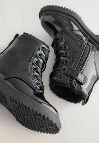 Schwarze Lackleder-Stiefeletten mit strukturiertem Obermaterial, Schnürung vorne und seitlichem Reißverschluss. Gefüttertes Innenleben mit Fell und gummierter Außensohle für besseren Halt.