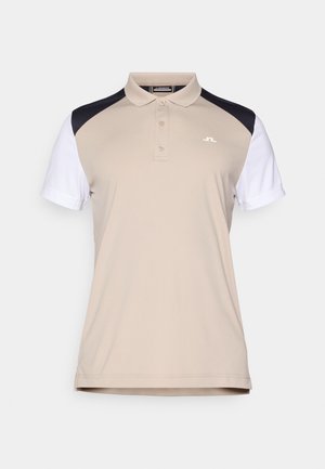 Beige pikétröja med korta vita ärmar och svarta axelpaneler. Den har en treknappars knäppning och en liten logotyp på bröstet.