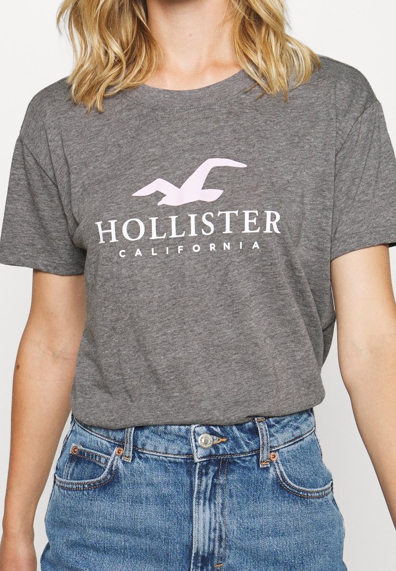 Grå bomullströja med korta ärmar, med en rosa måslogotyp och texten "HOLLISTER CALIFORNIA" i vitt nedanför. Enkel, avslappnad passform.