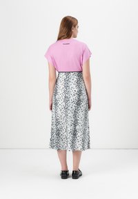 Une femme aux longs cheveux est vue de dos, portant un haut rose et une jupe à pois noir et blanc, se tenant devant un fond blanc uni.