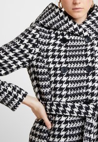 Cappotto in bianco e nero a motivo Houndstooth realizzato in tessuto testurizzato, con ampio colletto, design doppiopetto e cintura da annodare in vita.