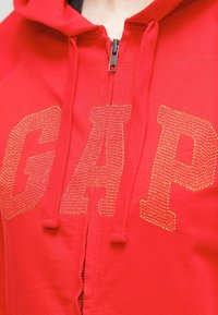 Czerwona bluza z kapturem na zamek błyskawiczny z dużym, teksturowanym złotym logo "GAP" z przodu. Posiada regulowany kaptur oraz miękki materiał bawełniany.