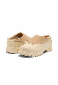 Chaussures beige à enfiler avec semelles épaisses et robustes, cols rembourrés et détails cousus, placées côte à côte sur un fond blanc.