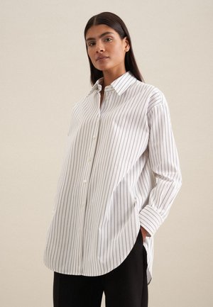 Witte button-up blouse met verticale zwarte strepen, lange mouwen en een gebogen zoom. De stof lijkt glad en licht glanzend.