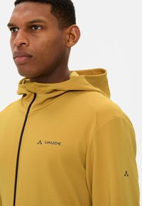 Gelbe Vaude Kapuzenjacke aus strukturiertem Stoff, mit Reißverschluss und Logodetail. Sie hat einen hohen Kragen und eine verstellbare Kapuze.