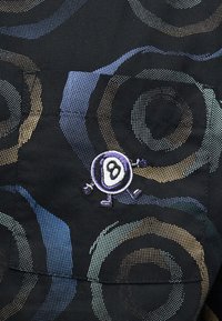 Juoda medžiaga su daugiamačių spalvotų taškuotų apskritimų raštais ir maža išsiuvinėta ėjimo aštuonių kamuoliukų aplikacija ant priekinių kišenių.