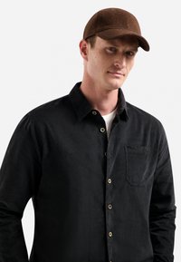 Zwarte corduroy overhemd met knopen, met een voorzak. Bruine pet met een gestructureerd oppervlak. Eenvoudig ontwerp, neutrale kleuren.