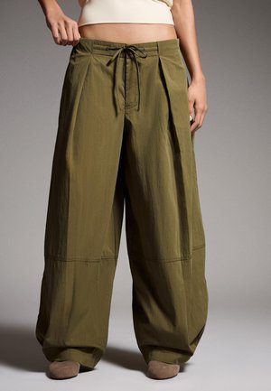 Pantaloni largi verde măsliniu, largi, cu talie ajustabilă prin șnur, purtați de o persoană care stă într-un decor simplu.