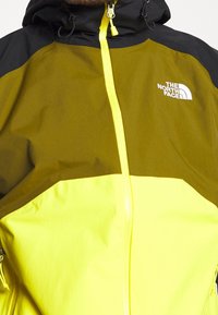 Giacca gialla e verde oliva con zip frontale, dettagli neri e cappuccio. Presenta un logo sul petto e hardware della zip visibile.