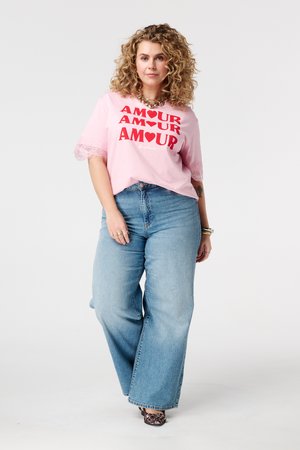 Femme aux cheveux bouclés portant un t-shirt rose "AMOUR", un jean à jambes larges bleu, des chaussures à motif léopard et des bijoux en or, se tenant avec assurance.
