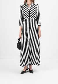 Robe longue à rayures noires et blanches avec fermeture par bouton, col et manches trois-quarts. Associée avec des chaussures à talons ouvertes noires et un sac à main.